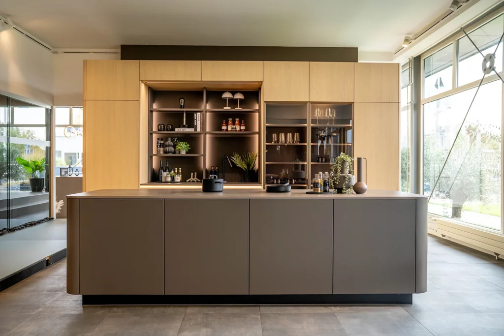 Moderne küchen mit insel wie in KOJE 25: KOCHINSEL in mattem taupe mit gerundeten enden, dahinter hochschränke in heller eiche mit beleuchteten offenen nischen und einer glasvitrine, im SHOWROOM in Weinheim